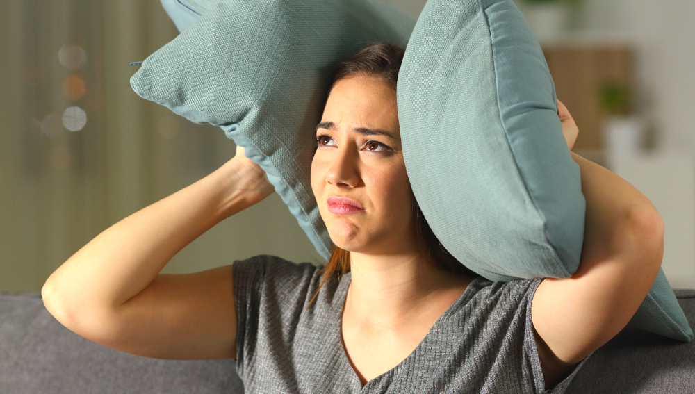 A distressed looking woman holding a pillow over her ear