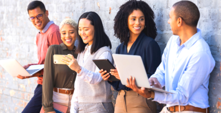 A group of smiling people standing nezt to a wall holding laptops, smart phones and tablets.