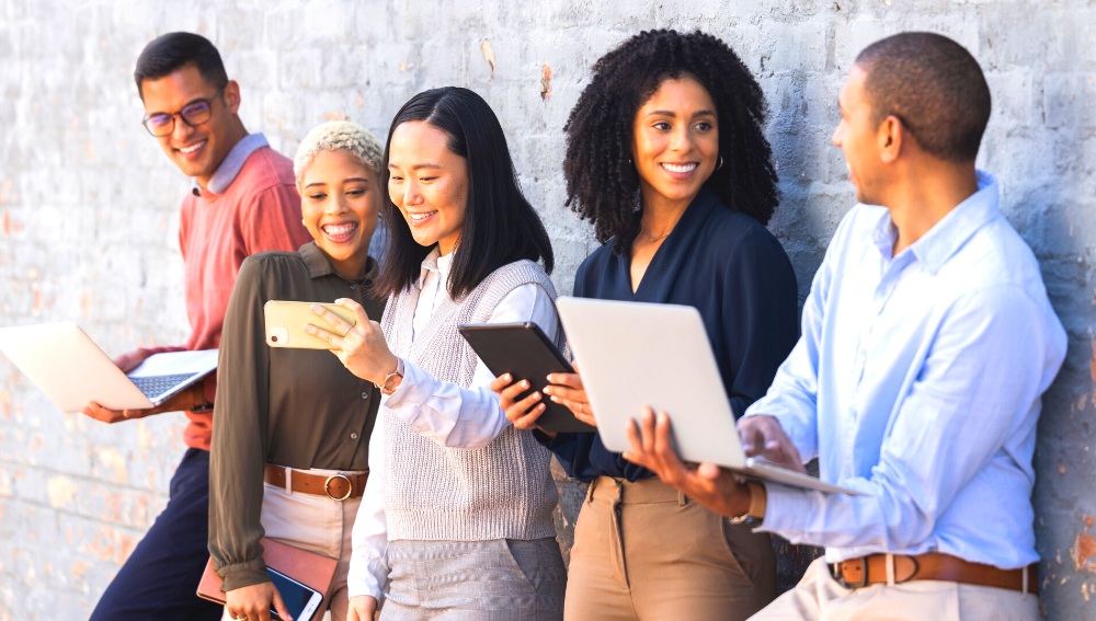 A group of smiling people standing nezt to a wall holding laptops, smart phones and tablets.