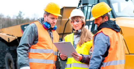 Two men and a woman wearing hard hats and safety vests in front of a TLB