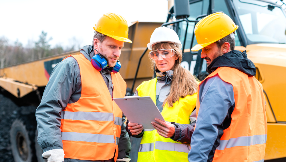 Two men and a woman wearing hard hats and safety vests in front of a TLB